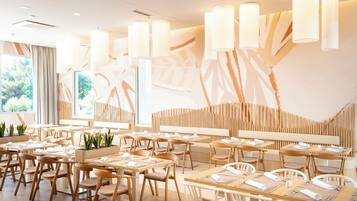 A modern restaurant interior with wooden furniture, pendant lighting, and a view of greenery outside.