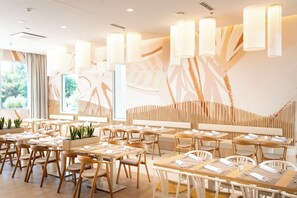 Un intérieur de restaurant moderne, avec des meubles en bois, des luminaires suspendus et une vue sur la verdure à l’extérieur.