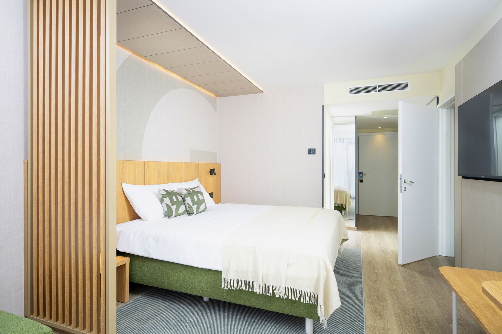 A hotel room with a bed, a wooden headboard, a green upholstered bed base, a white blanket, and a TV on the wall.
