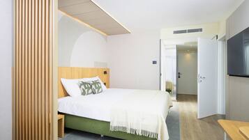 A hotel room with a bed, a wooden headboard, a green upholstered bed base, a white blanket, and a TV on the wall.