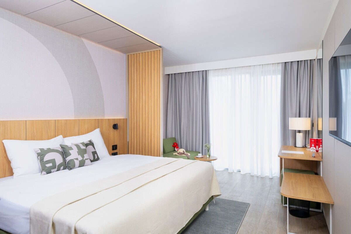 Une chambre d’hôtel moderne avec un grand lit, un bureau avec une lampe et une vue sur la ville grâce à de grandes fenêtres.