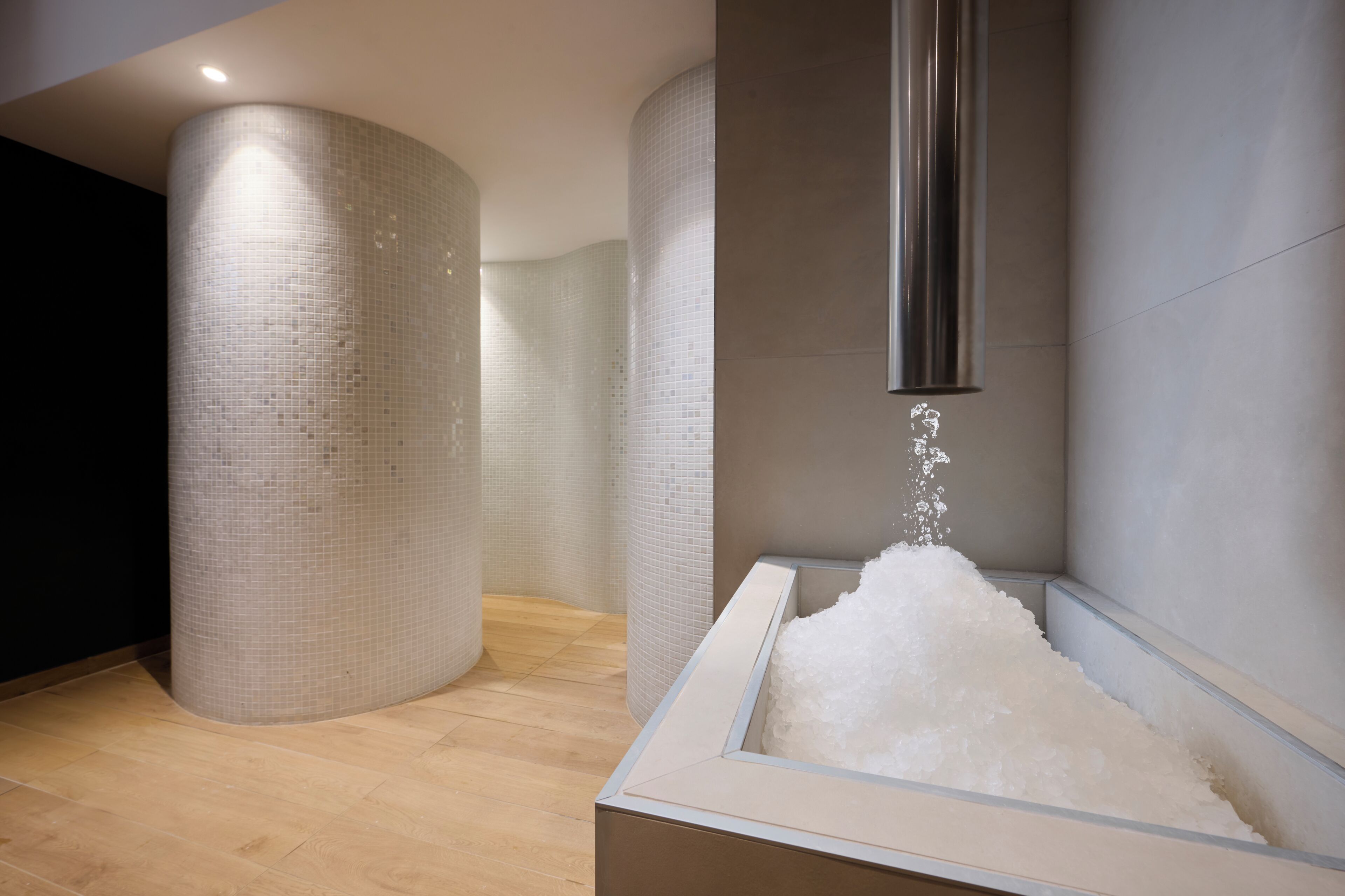 Een moderne badkamer met een grote, cilindrische kolom, een douchegedeelte waar water in een rechthoekig wasbak stroomt, en een houten vloer.
