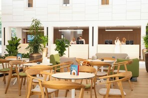 Un hall d’hôtel avec des tables et des chaises rondes, un comptoir d’accueil et des plantes en pot.