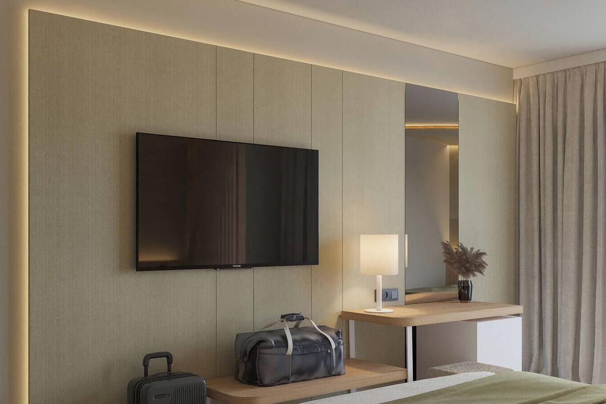 Een hotelkamer met een flatscreen televisie aan de muur, een nachtkastje met een lamp en een vaas gedroogde bloemen, een koffer en een bed met een groene deken.