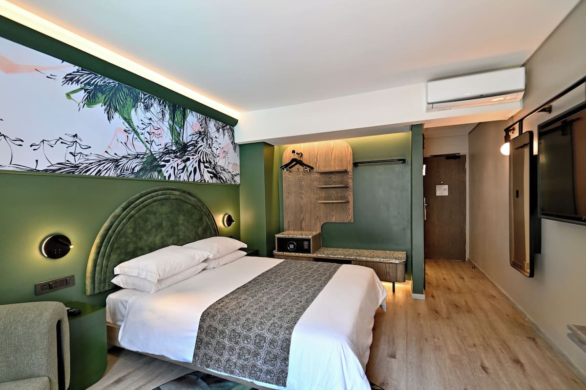 Una habitación de hotel moderna con una cama grande, un cabecero con iluminación integrada, un televisor montado en la pared y una pintura de follaje tropical.