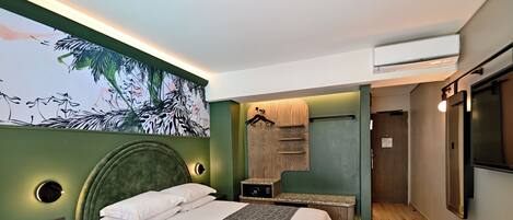 Una habitación de hotel moderna con una cama grande, un cabecero con iluminación integrada, un televisor montado en la pared y una pintura de follaje tropical.