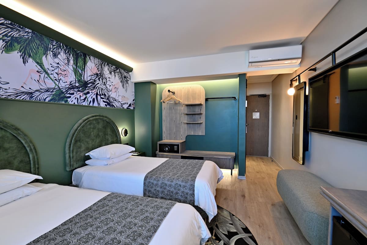 Habitación de hotel con dos camas, un escritorio, una silla, un televisor, aire acondicionado y un perchero en la pared.