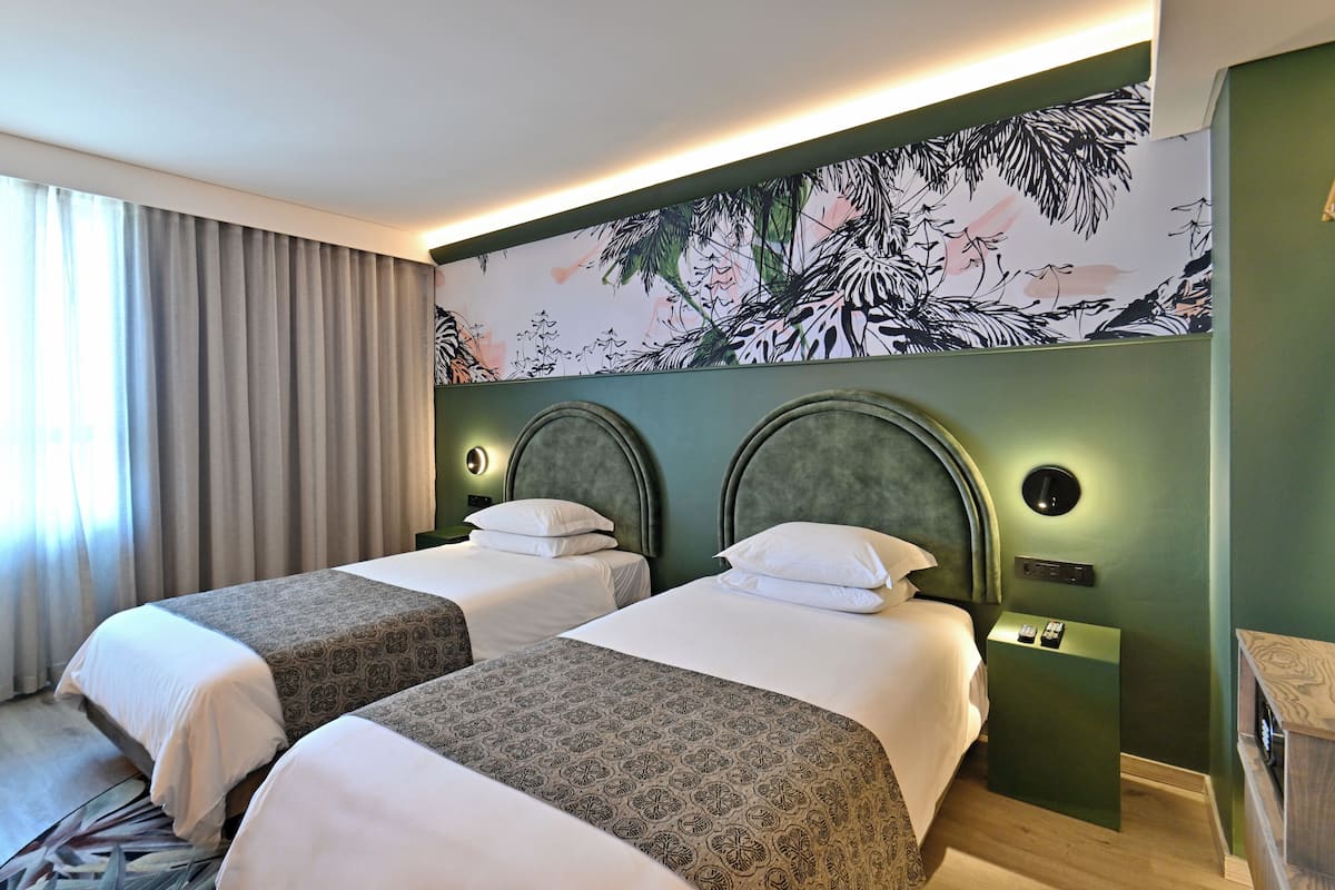 Habitación de hotel con dos camas, un ventanal con cortinas, un mural en la pared y una mesita con lámpara.