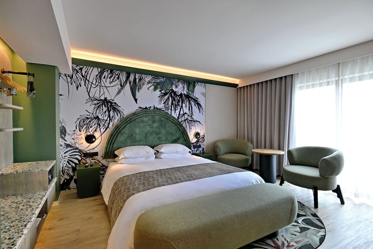 Habitación de hotel con cama, un sillón tapizado en verde, una mesa redonda y una pared con estampado de hojas tropicales.