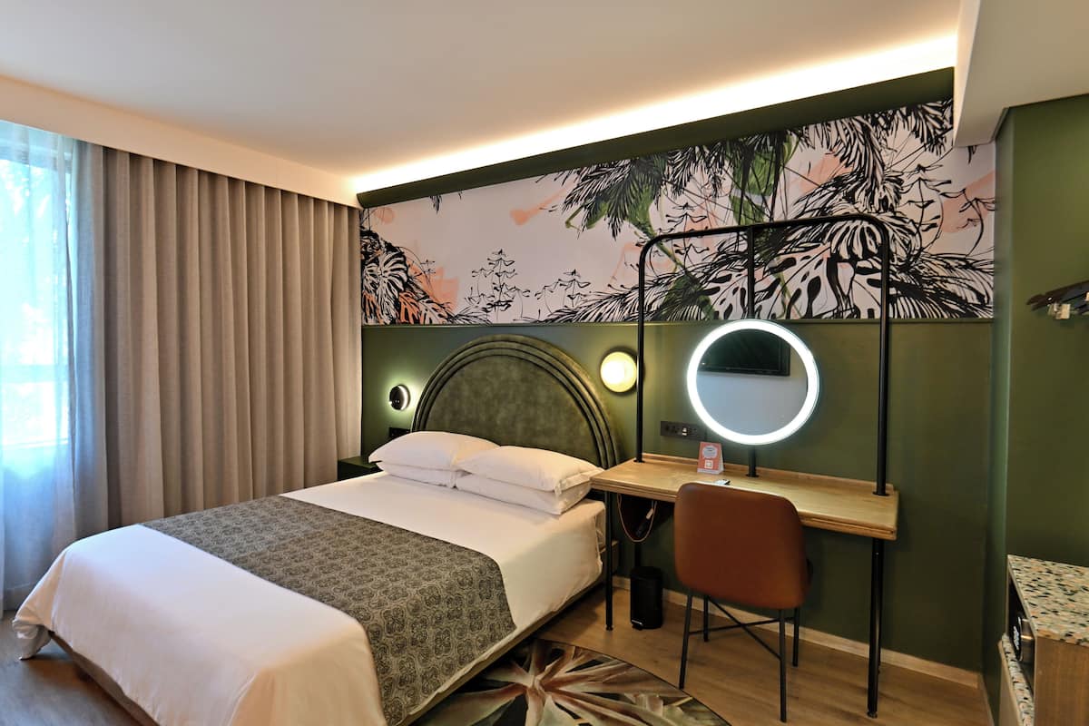 Habitación de hotel con cama, escritorio, silla, espejo redondo y pared con diseño de hojas.