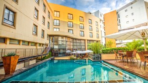 A swimming pool with a glass enclosure, surrounded by outdoor seating and a building with multiple balconies. - City Lodge Hotel Umhlanga Ridge (Umhlanga)