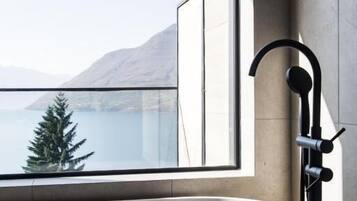 Un baño moderno con una bañera grande, un ventanal con vistas a un lago y montañas, y un cabezal de ducha manual negro.
