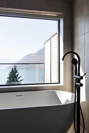 A modern bathroom with a large bathtub, a window with a view of a lake and mountains, and a black handheld showerhead.