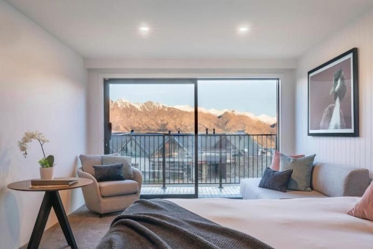 A bedroom with a large bed, a chair, a small table, and a balcony with a view of mountains.