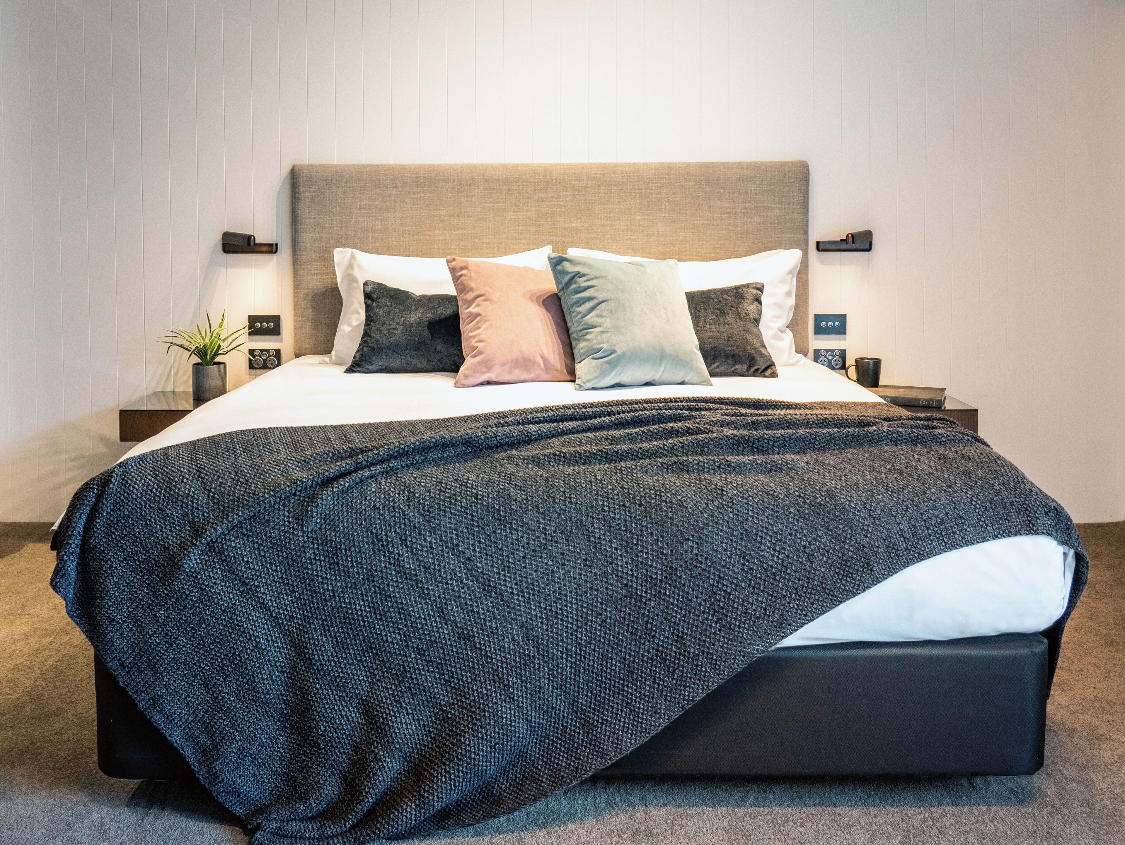 A neatly made bed with a dark blanket, multiple pillows, bedside tables with lamps, and a potted plant.