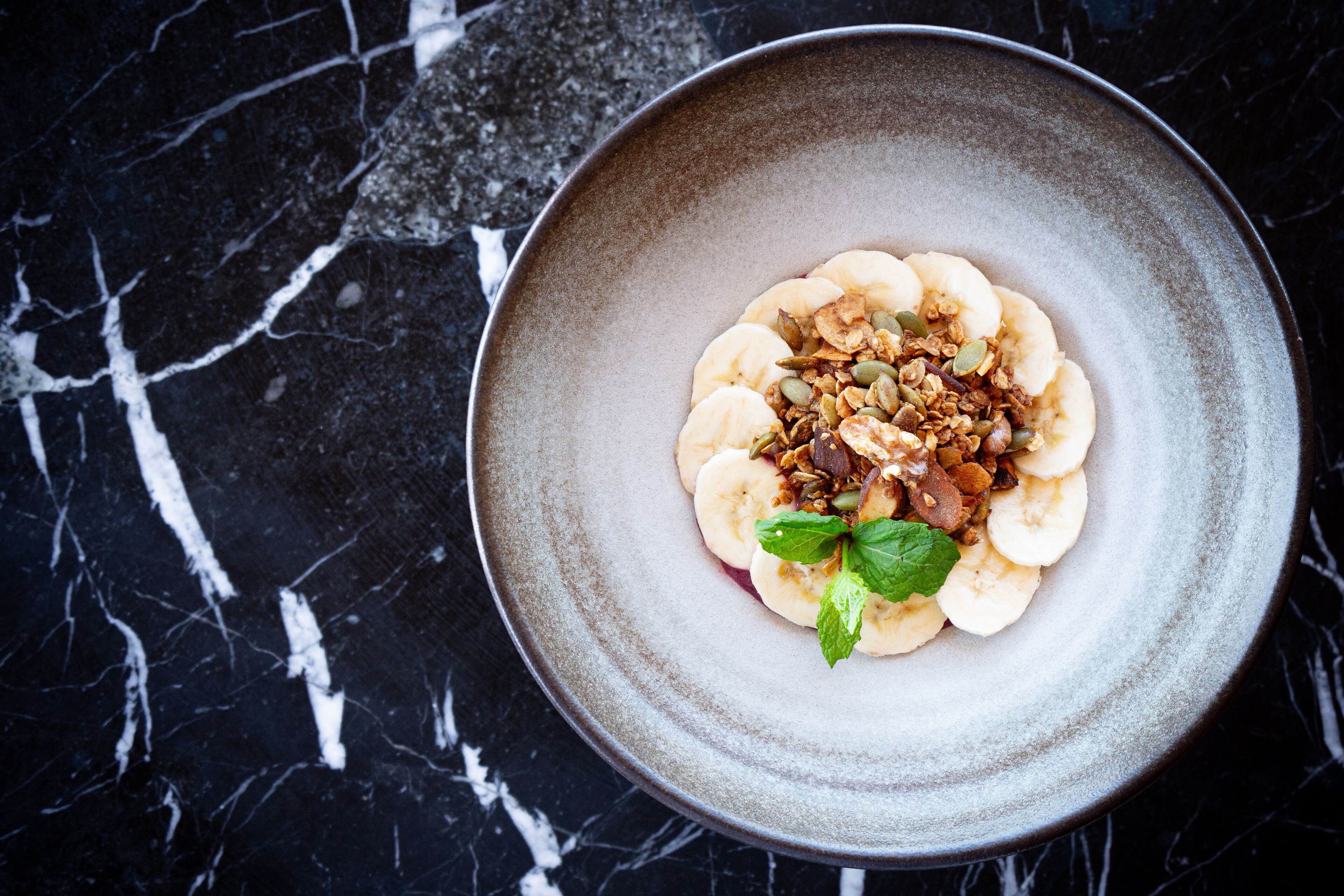 A plate of food with bananas, granola, and a mint leaf on a dark marble surface.
