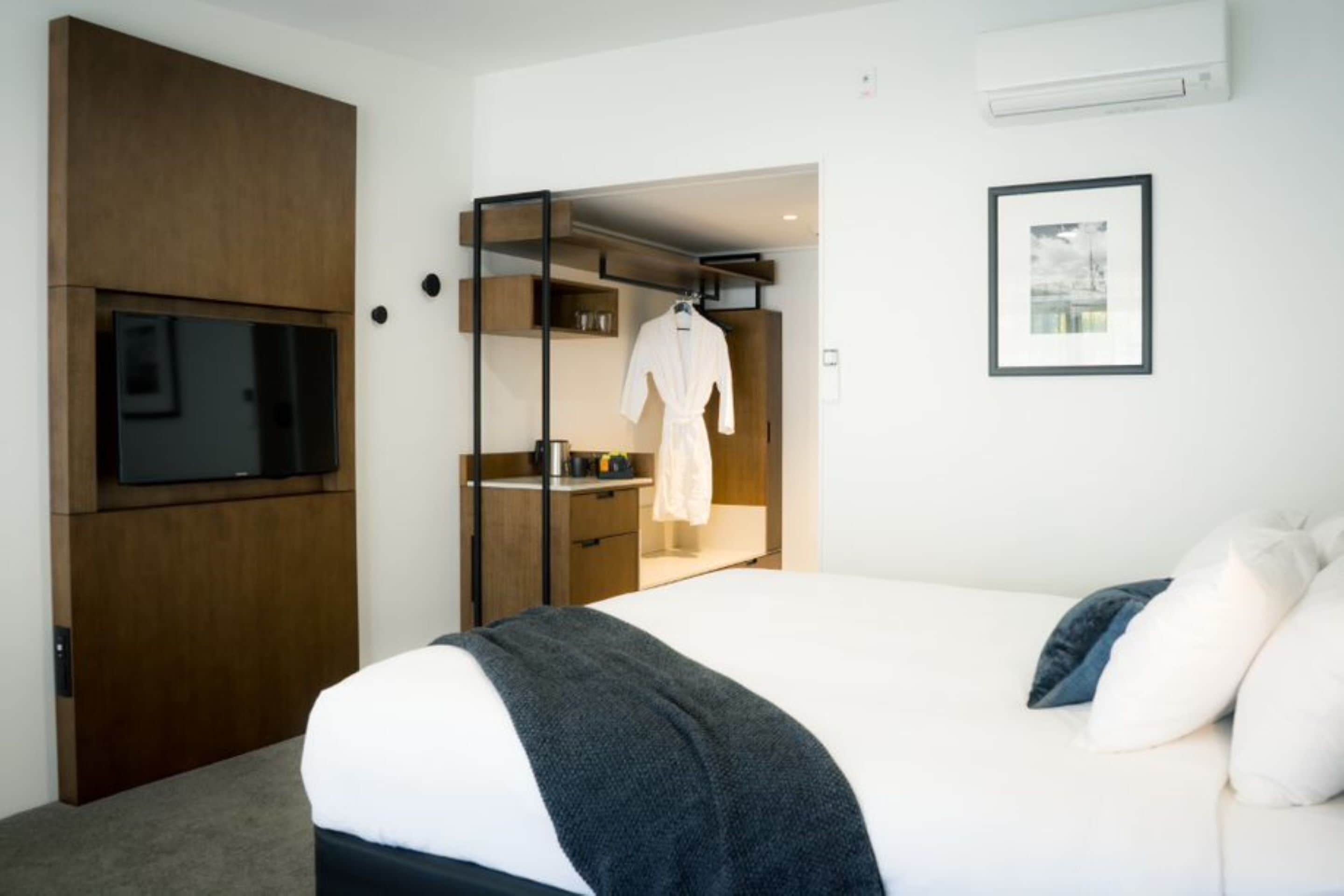 A modern hotel room with a large bed, a TV mounted on a wooden cabinet, a wardrobe, and a framed picture on the wall.