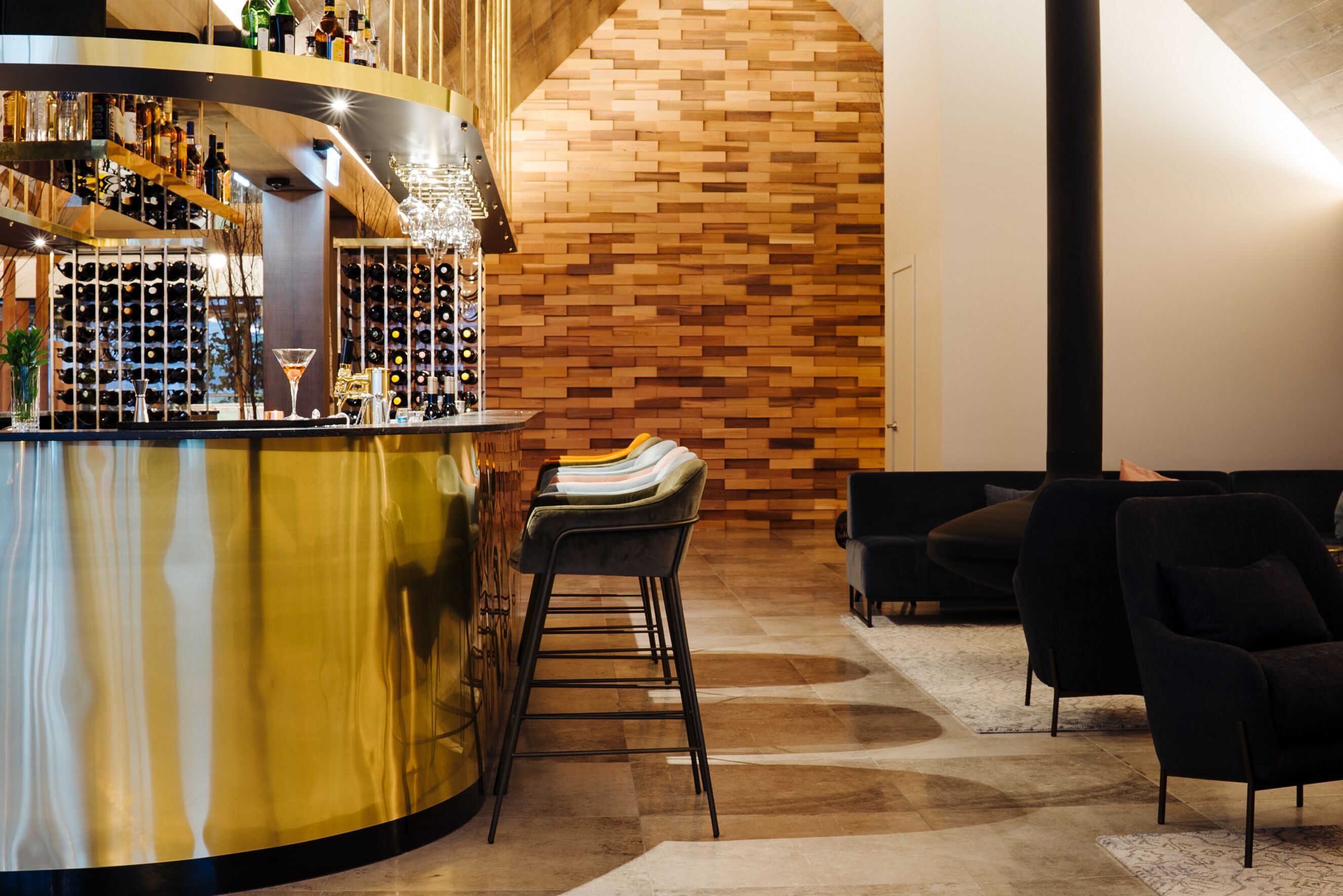 A bar area with a curved counter, multiple bottles, and a wine rack. There are three bar stools and two black chairs in the seating area.