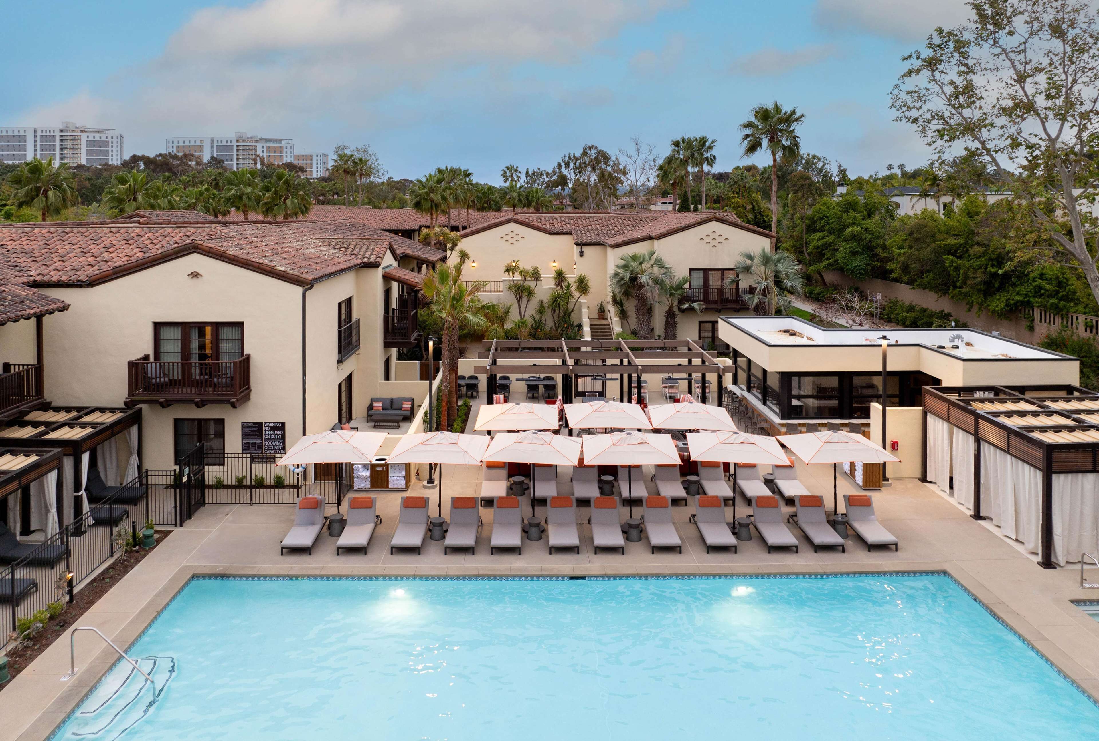 A pool area with lounge chairs and umbrellas, surrounded by buildings with balconies and a view of palm trees.