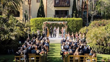An outdoor wedding ceremony with guests seated on wooden chairs arranged in rows, facing a stage with a floral arch.