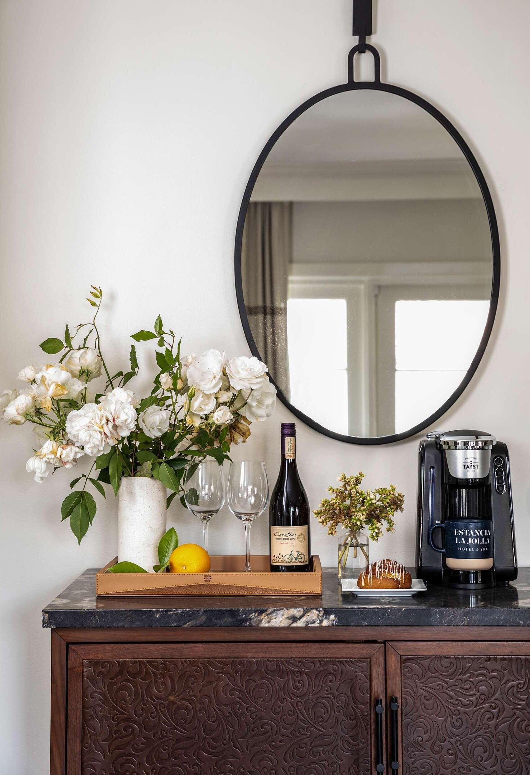 Una encimera de cocina con un florero de flores blancas, dos copas de vino, una botella de vino, una planta pequeña y una cafetera.