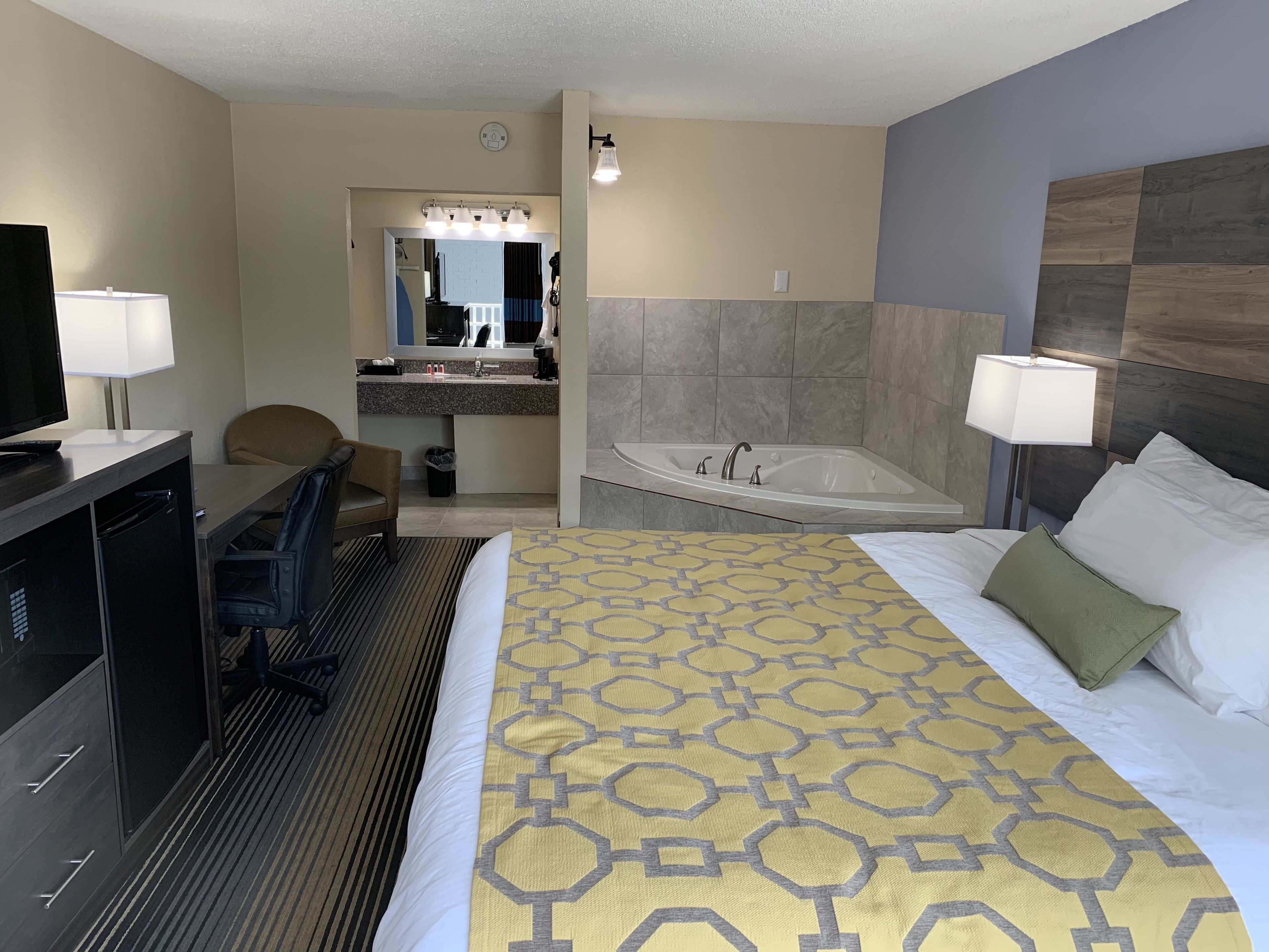 A hotel room with a bed, a desk with a chair, a TV, a bathroom with a bathtub, and a mirror.