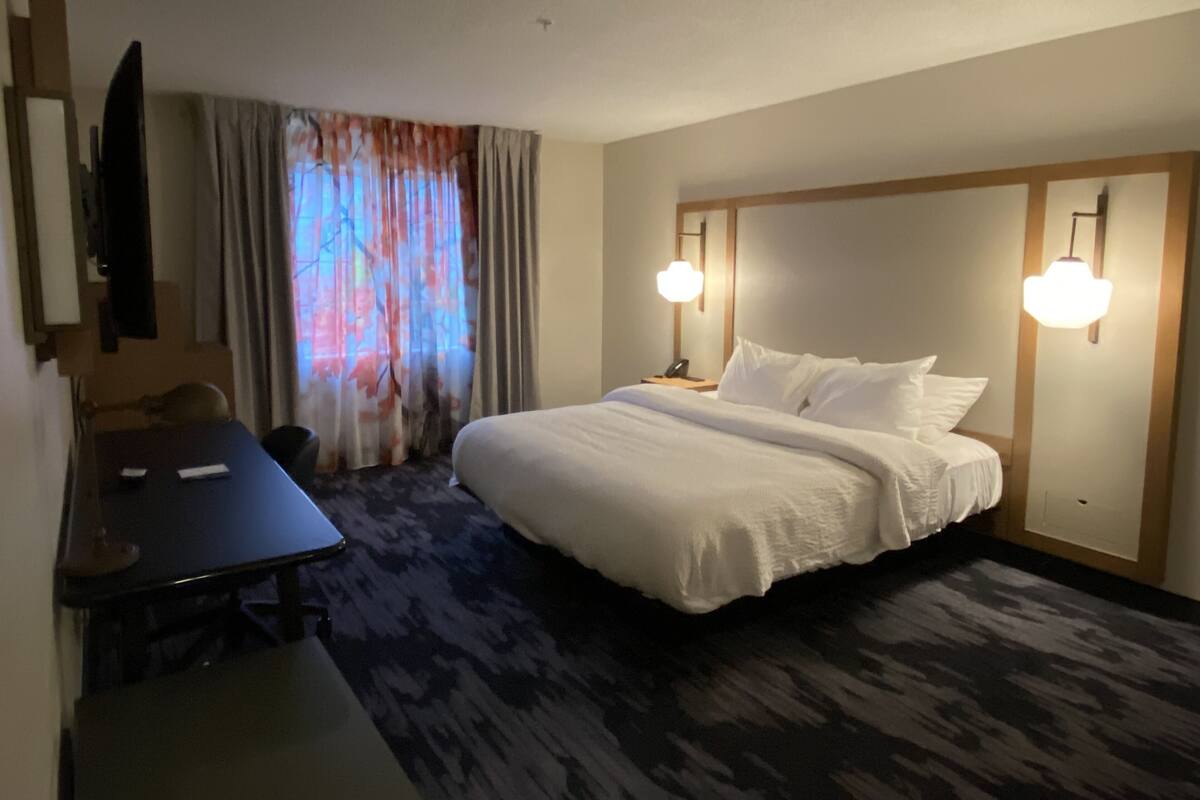 Habitación de hotel con cama, lámparas de noche, escritorio, silla, televisor y ventana con cortinas.