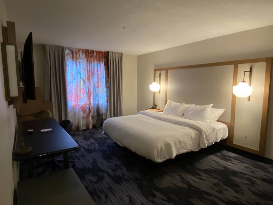 A hotel room with a bed, bedside lamps, a desk, a chair, a TV, and a window with curtains.