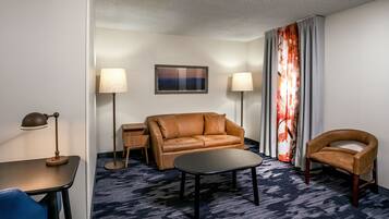 Una habitación de hotel con un sofá de cuero marrón, una mesita negra pequeña, una silla azul, dos lámparas de pie y una alfombra estampada.