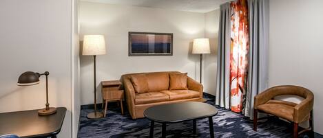 Una habitación de hotel con un sofá de cuero marrón, una mesita negra pequeña, una silla azul, dos lámparas de pie y una alfombra estampada.