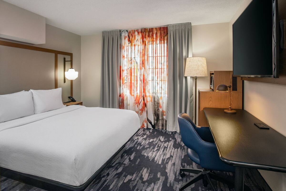 Habitación de hotel con una cama grande, un escritorio, una silla, un televisor y una ventana con cortinas.