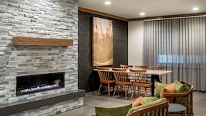 A modern living room with a stone fireplace, wooden dining table, and green upholstered chairs. - Fairfield Inn & Suites by Marriott Saratoga Malta (Ballston Spa)
