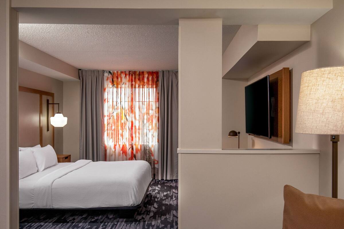 Habitación de hotel con cama, televisión montada en la pared, lámpara y ventana con cortinas.