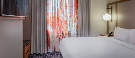 A hotel room with a large bed, a TV on a stand, a bedside table with a lamp, and a window with sheer curtains.