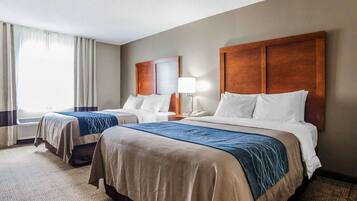A hotel room with two beds, a large wooden headboard, a bedside table with a lamp, and a view of the outdoors.
