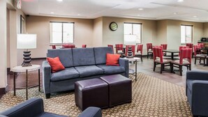 Un hall d’hôtel avec un canapé gris, des coussins rouges, une horloge et un coin repas avec des chaises et des tables rouges.