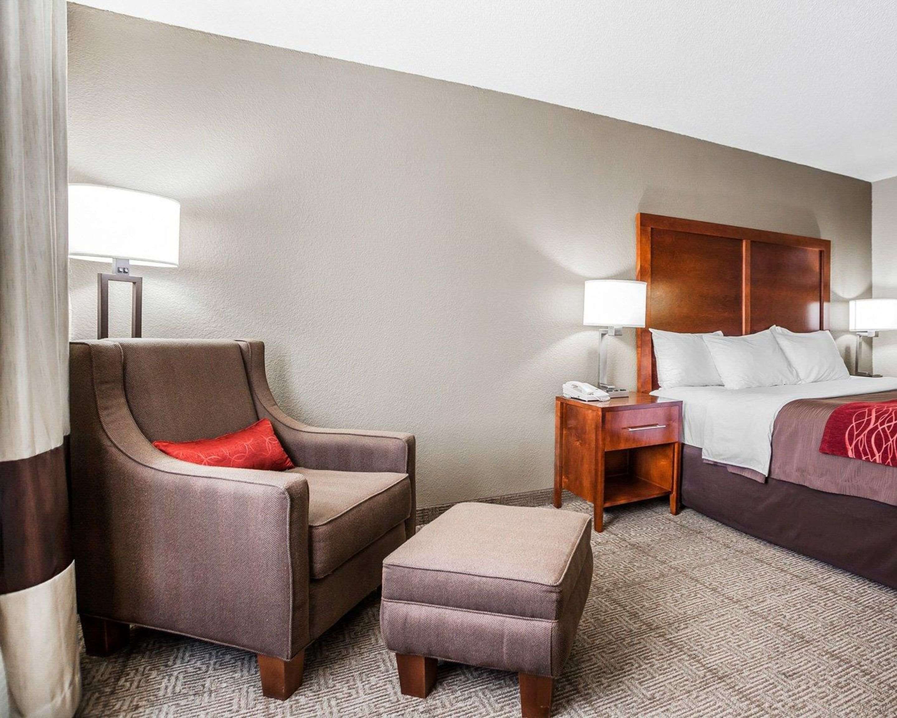 A hotel room with a bed, a chair, an ottoman, a nightstand, and a lamp.