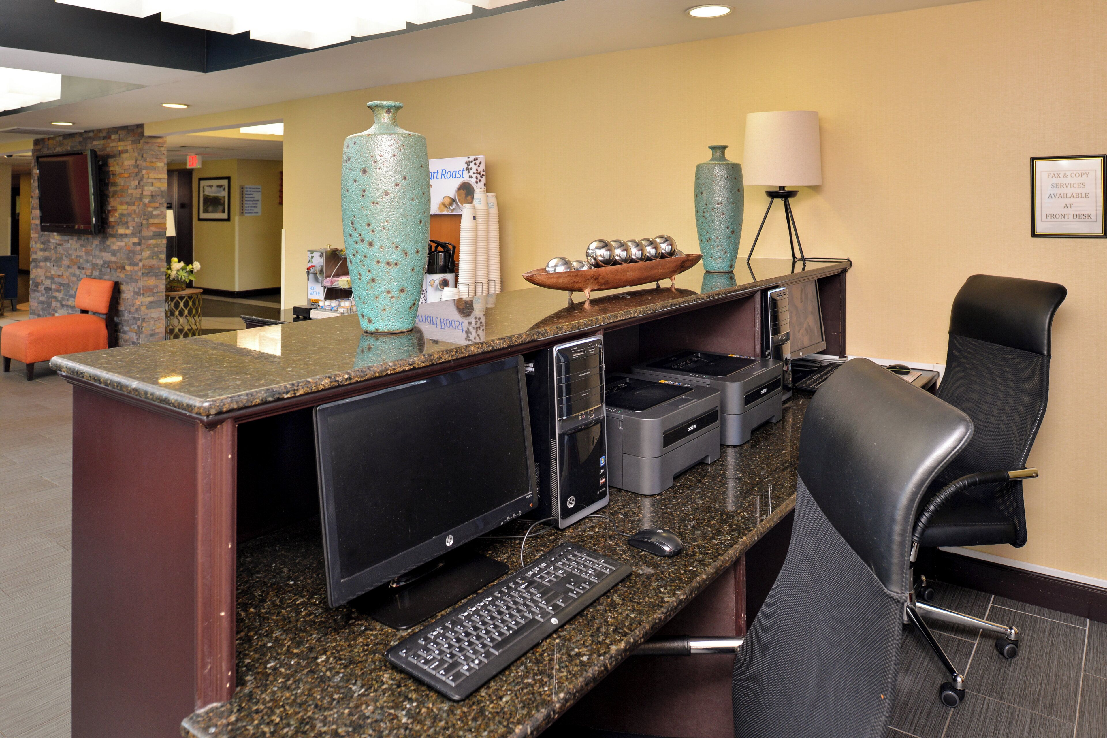 A reception desk with a computer, printer, and keyboard. There is a chair and a lamp on the desk.