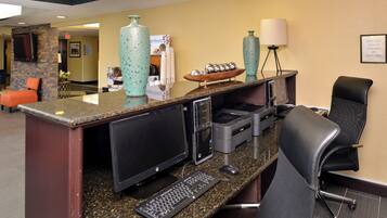 A reception desk with a computer, printer, and keyboard. There is a chair and a lamp on the desk.