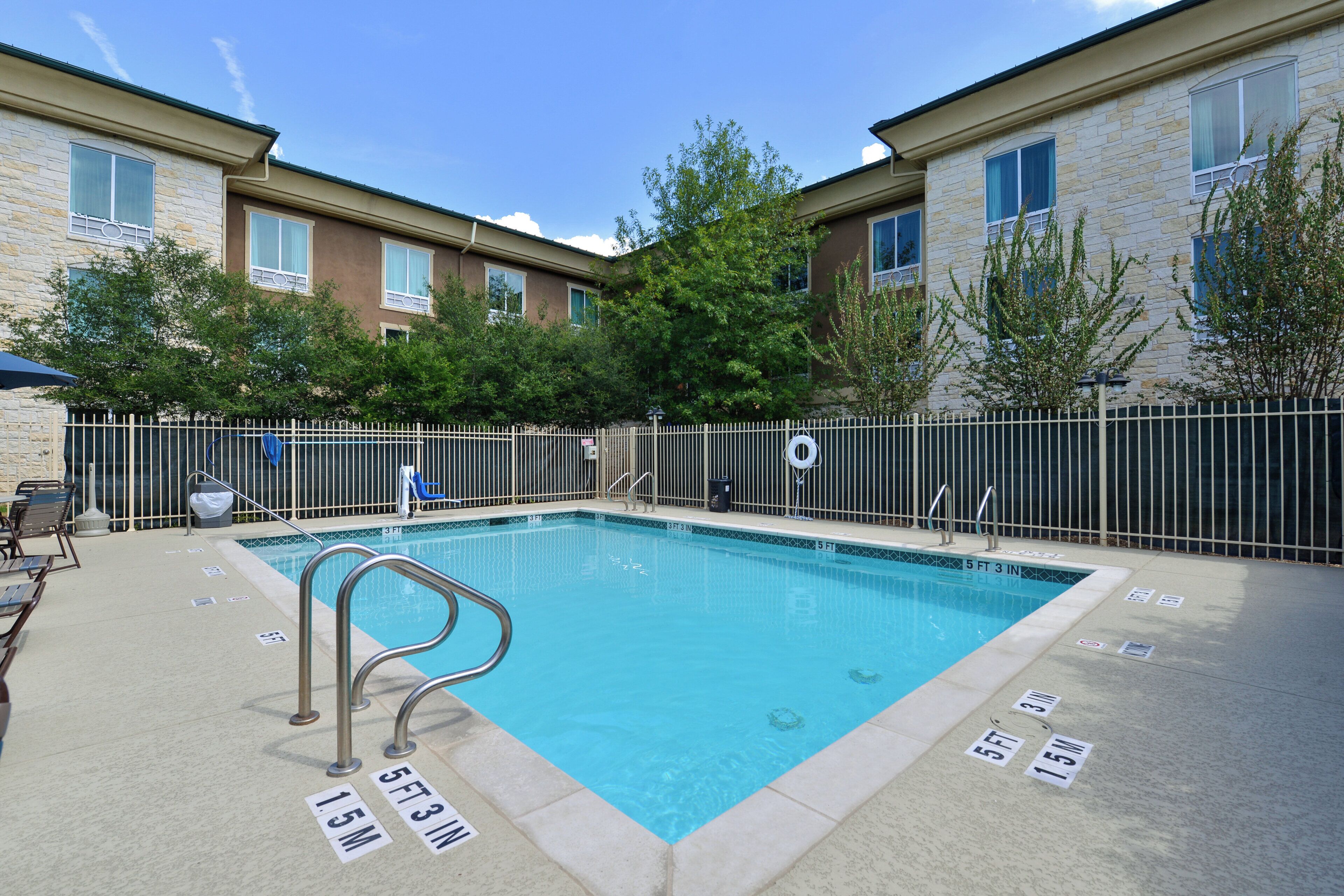 A rectangular pool with a metal ladder, surrounded by a fence, and adjacent to a building with multiple windows and a brick facade.