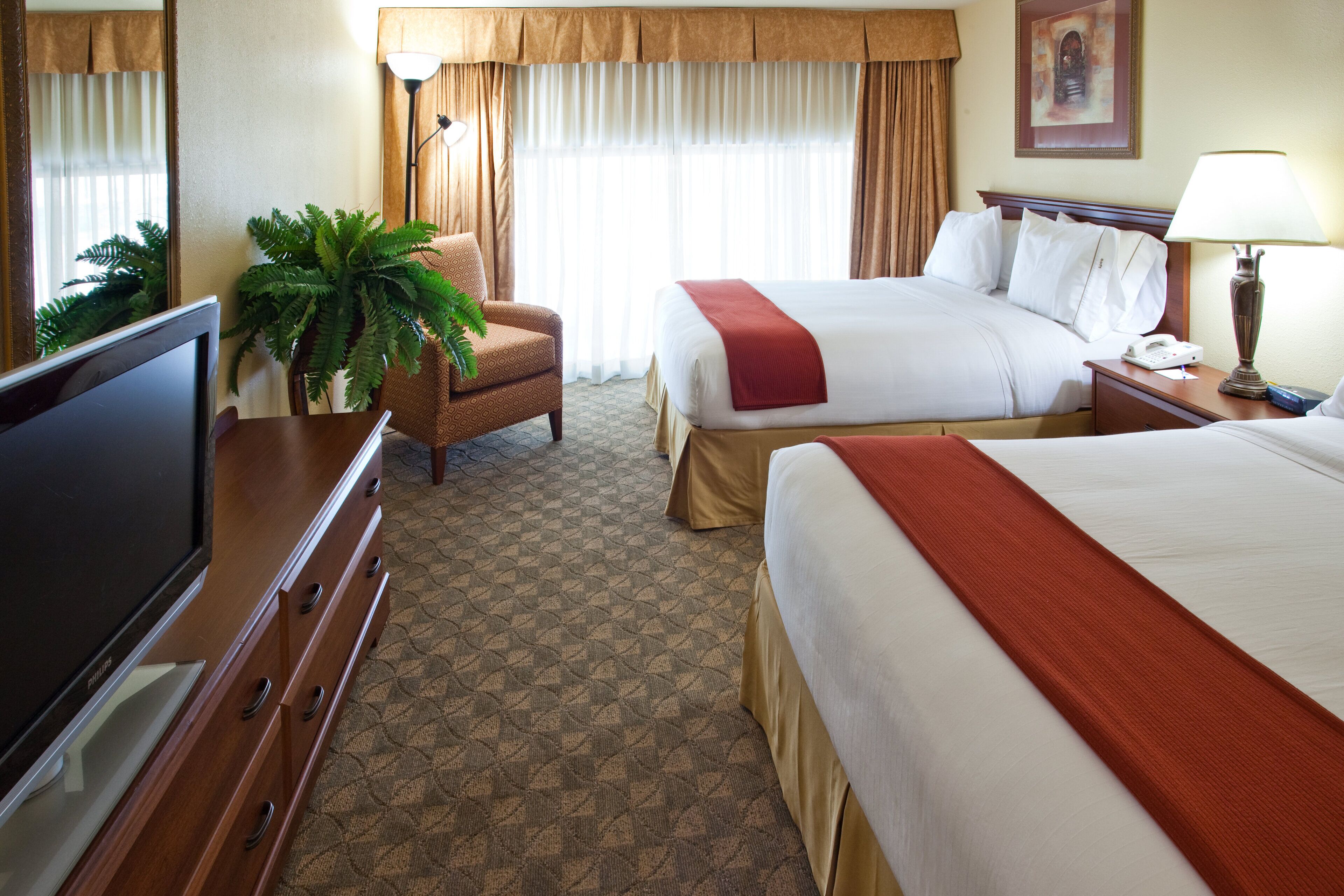 A hotel room with a large bed, a smaller bed, a desk with a TV, a chair, a lamp, and a plant.