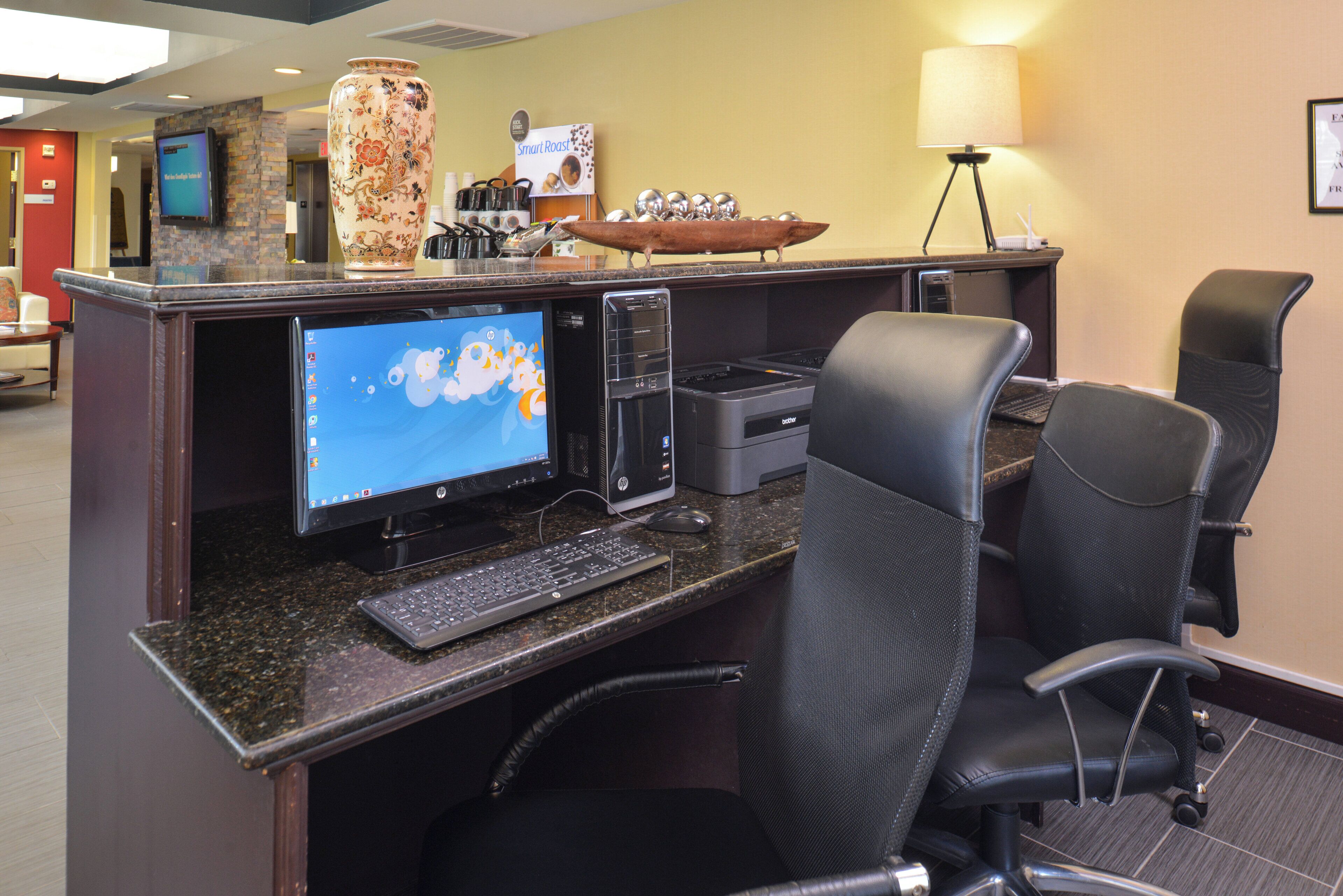 A hotel reception area with a desktop computer, two chairs, a coffee station, and a lamp.