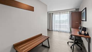 A modern hotel room with a wooden bench, a desk with a computer, and a large window with sheer curtains.