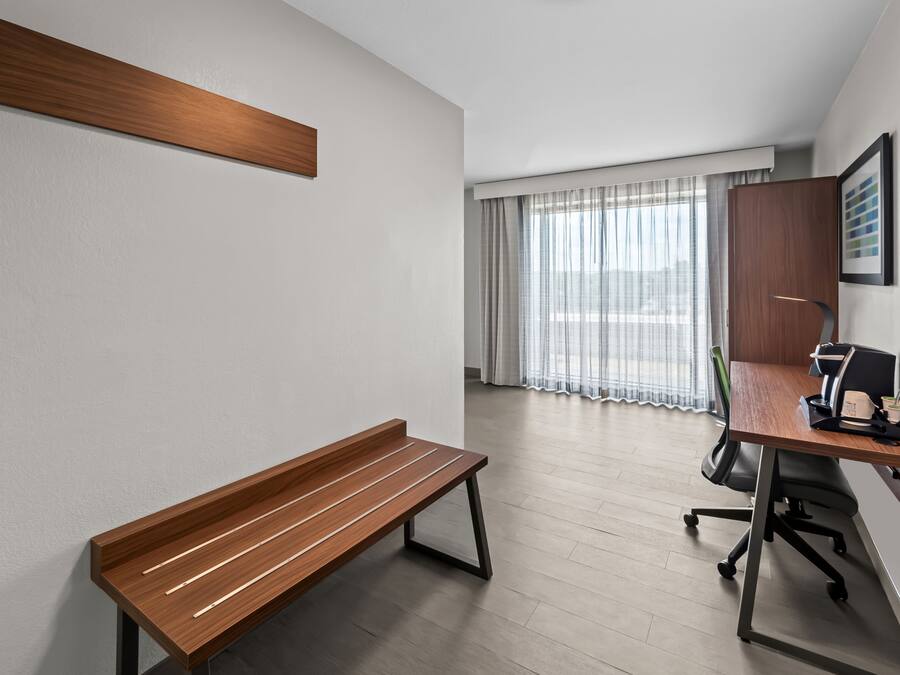 A modern hotel room with a wooden bench, a desk with a computer, and a large window with sheer curtains.