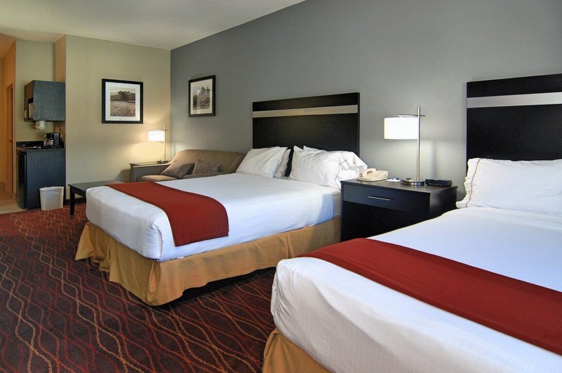 A hotel room with two beds, a desk, a chair, a lamp, and a small kitchenette.