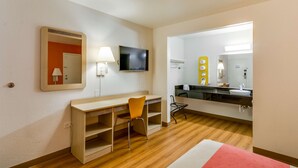 A hotel room with a wooden desk, a chair, a television, a mirror, and a bathroom with a sink and mirror. - Motel 6 Schiller Park, IL - Chicago O'Hare (Schiller Park)