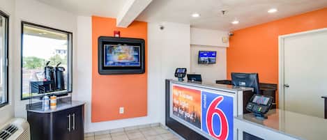 Zona de recepción de motel con una pantalla digital, mostrador con un letrero con el número '6' y ventana con vistas al exterior.
