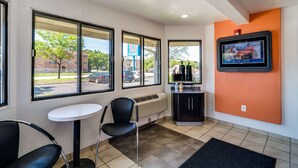 A hotel lobby with large windows, a small table, chairs, a radiator, and a television mounted on the wall. - Motel 6 Schiller Park, IL - Chicago O'Hare (Schiller Park)
