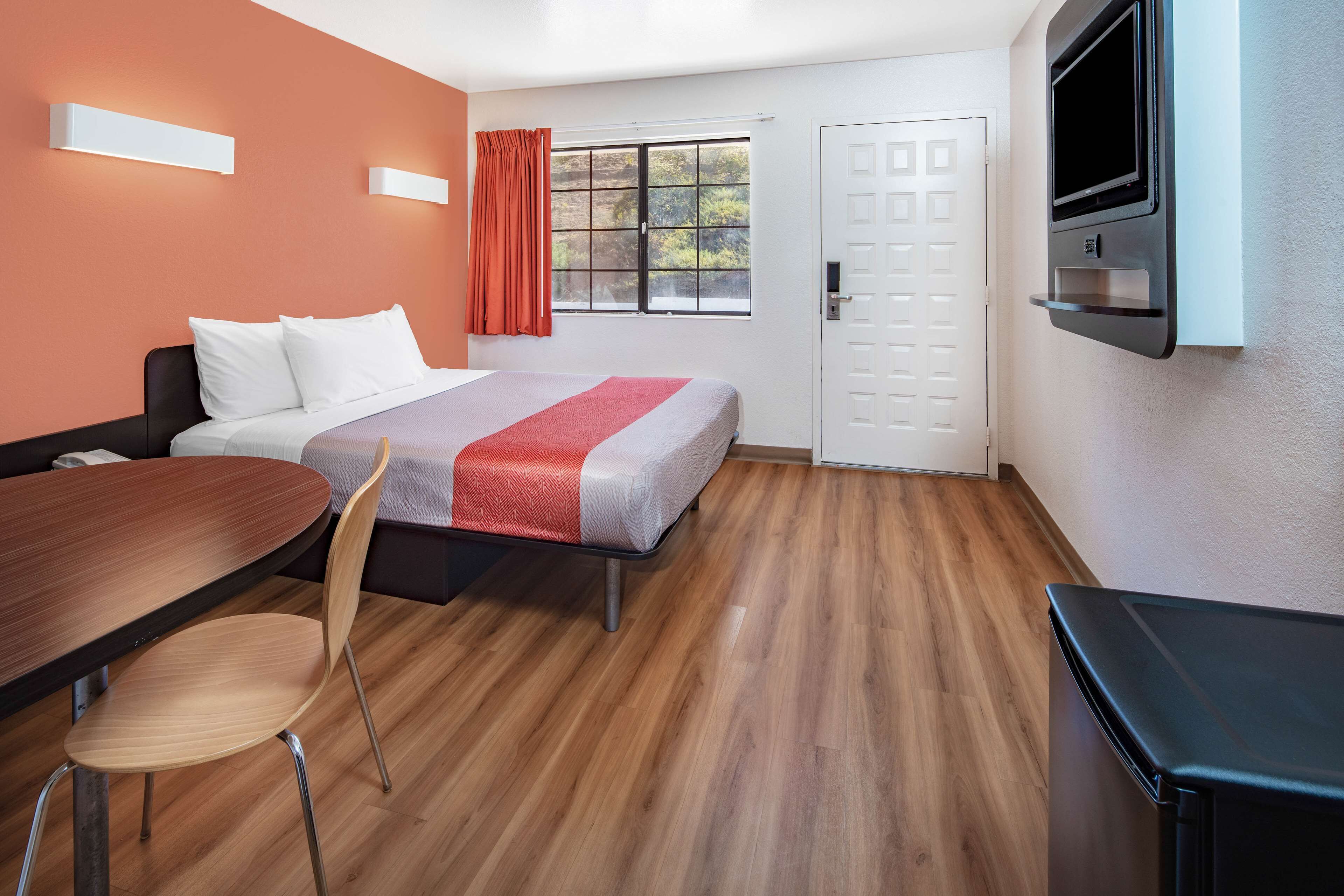 A hotel room with a bed, a TV mounted on the wall, a dining table with chairs, and a window with curtains.