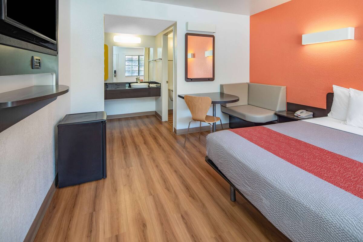 Une chambre d’hôtel avec un lit, un bureau, une chaise, un petit réfrigérateur et une salle de bain visible à travers une porte ouverte.